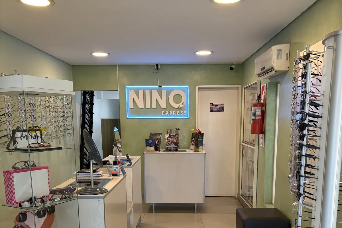 El local por dentro: los exhibidores de anteojos a los costados y, al fondo, el cartel de Nino Express iluminado en azul sobre la pared verde